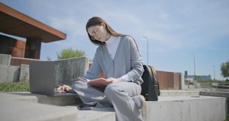 A hand-held shot of a young female student in casual clothes and glasses using a laptop and taking notes with a pen in a notebook in an urban park. © Anatoly Repin