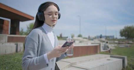 A hand-held shot of a young female student in casual clothes, wearing glasses and headphones, sitting on a concrete bench in an urban park, listening to music and drinking water from a bottle. © Anatoly Repin