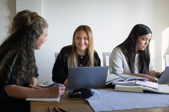 Smiling blond teenage girl talking with mother while studying with friend at home
