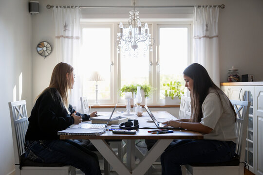 Multiracial teenage girls doing homework on laptop while sitting at table in living room