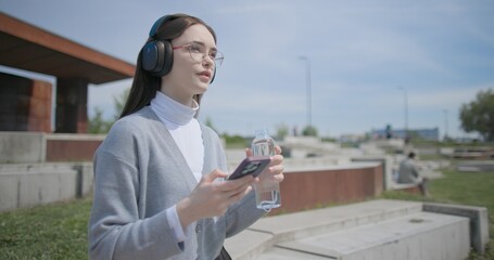 A hand-held shot of a young female student in casual clothes, wearing glasses and headphones, sitting on a concrete bench in an urban park, listening to music and drinking water from a bottle. © Anatoly Repin