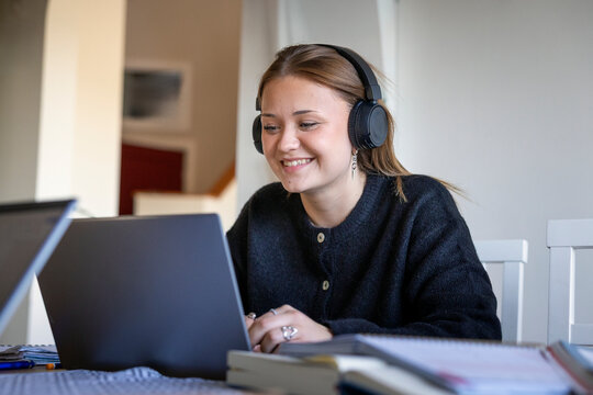 Smiling teenage girl wearing wireless headphones attending online lecture through laptop at home