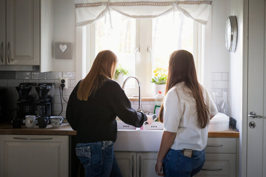 Rear view of teenage girl standing beside female friend washing dishes in kitchen at home