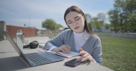 A hand-held shot of a young female student in casual clothes and glasses using a laptop and taking notes with a pen in a notebook in an urban park. © Anatoly Repin