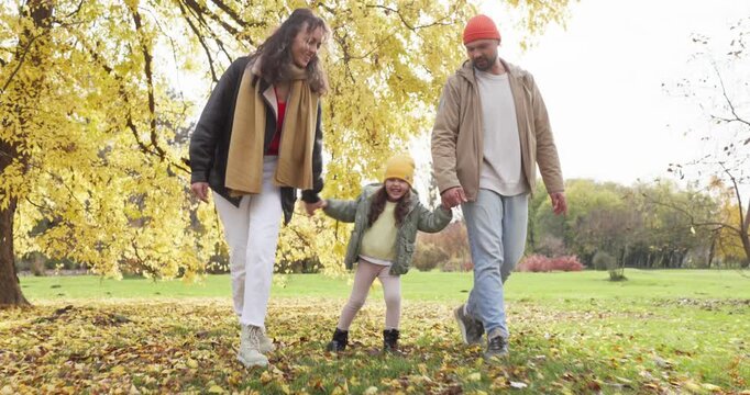 Happy family mother father daughter walking autumn park. Parents hold their child by the hands among yellow leaves and enjoy fresh air. Warm joyful bonding family concept for advertising.