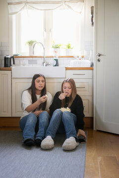 Full length of teenage girls eating popcorn while sitting on kitchen floor at home