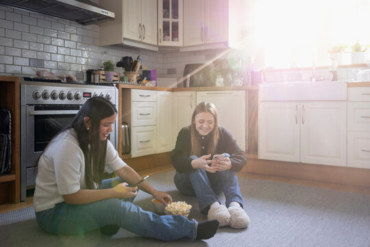 Teenage girls using smart phone and eating popcorn while sitting on floor in kitchen at home
