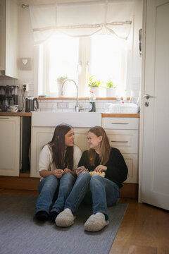Multiracial female friends eating popcorn while enjoying leisure time sitting on kitchen floor at home