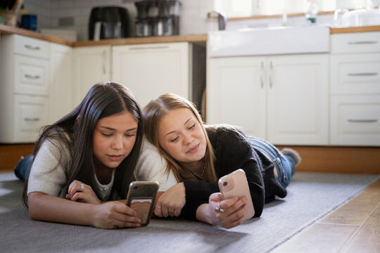 Carefree teenage girl lying beside female friend on kitchen floor and using using smart phone at home