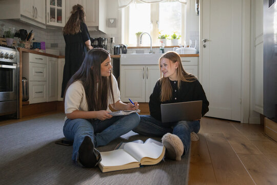 Smiling high school teenage girls studying with each other while sitting on kitchen floor at home