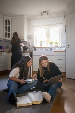 Full length of female friends studying with each other while sitting on kitchen floor at home