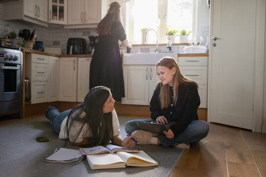 Teenage girl lying on kitchen floor while doing homework with friend at home