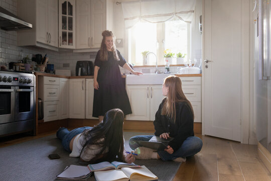 Mother talking with daughter doing homework with female friend while sitting on kitchen floor at home