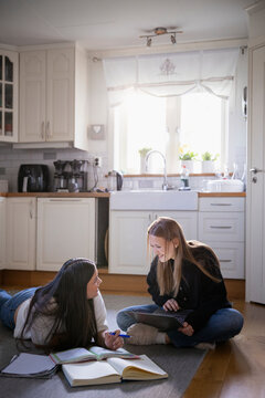 Smiling teenage girl talking with female friend lying on kitchen floor while doing homework in home
