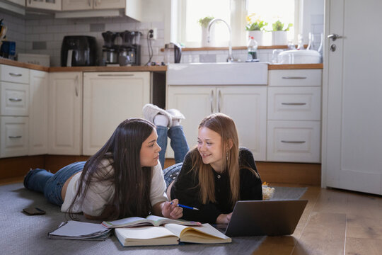 Multiracial female friends lying down on kitchen floor while doing homework at home