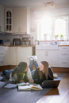 Smiling multiracial female friends lying down on kitchen floor while doing homework at home