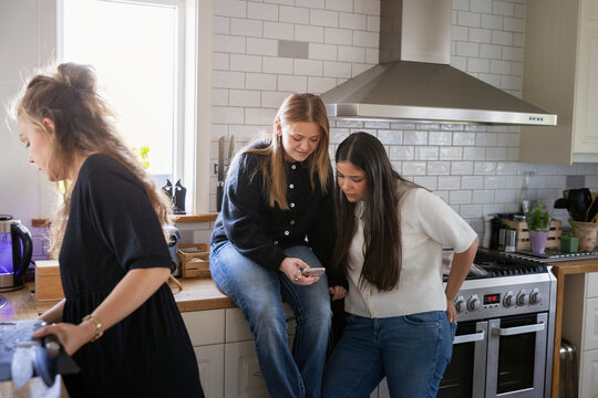 Female teenage friends surfing on smart phone while spending leisure time in kitchen at home