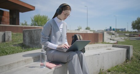 A hand-held shot of a young female student in casual clothes, glasses, and headphones working and typing on a laptop while sitting on a concrete bench in an urban park. © Anatoly Repin