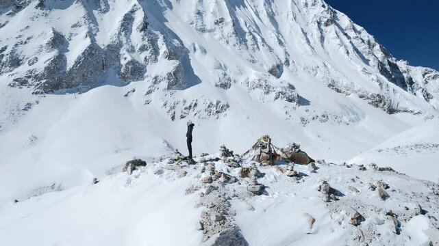 Drone shot of a trekker standing with arms raised in triumph on a snowy Himalayan ridge with prayer flags and cairns in Nepal's Manaslu region.