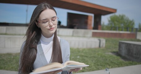 A hand-held shot of a young female student in casual clothes wearing glasses sitting on a concrete bench in an urban park on a sunny summer day and reading a book, flipping through the pages. © Anatoly Repin