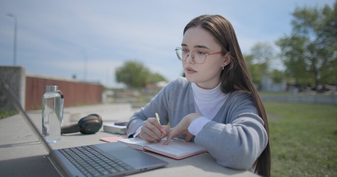 A hand-held shot of a young female student in casual clothes and glasses using a laptop and taking notes with a pen in a notebook in an urban park.
