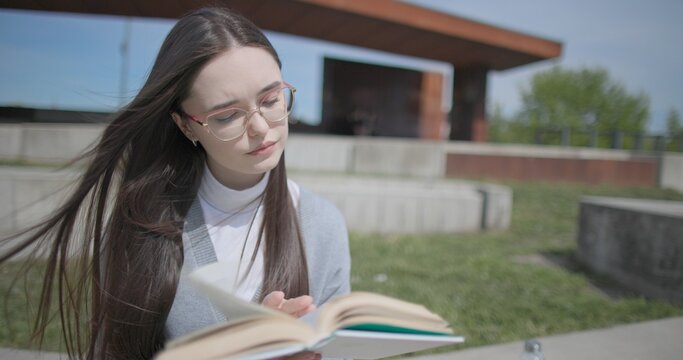 A hand-held shot of a young female student in casual clothes wearing glasses sitting on a concrete bench in an urban park on a sunny summer day and reading a book, flipping through the pages.