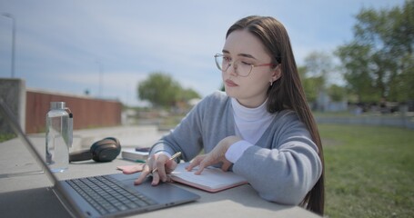 A hand-held shot of a young female student in casual clothes and glasses using a laptop and taking notes with a pen in a notebook in an urban park. © Anatoly Repin