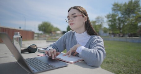 A hand-held shot of a young female student in casual clothes and glasses using a laptop and taking notes with a pen in a notebook in an urban park. © Anatoly Repin