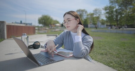 A hand-held shot of a young female student in casual clothes and glasses using a laptop and taking notes with a pen in a notebook in an urban park. © Anatoly Repin
