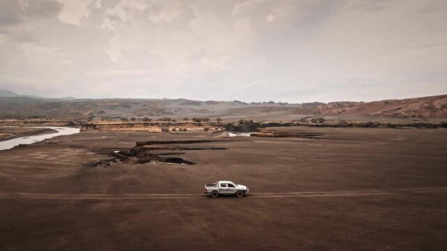 White pickup truck car driving volcanic landscape near Port Resolution on Tanna Island, dark ash, sand with eroded earth formations and a river flowing under overcast sky. Aerial view drone flight