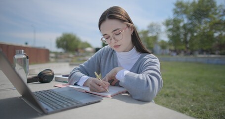 A hand-held shot of a young female student in casual clothes and glasses using a laptop and taking notes with a pen in a notebook in an urban park. © Anatoly Repin