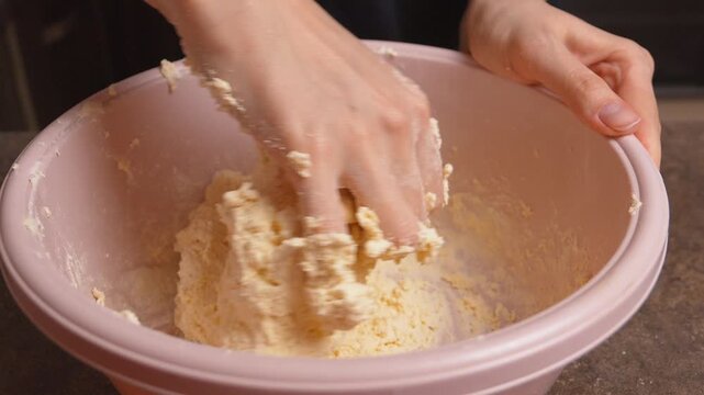 Female hands carefully kneading cottage cheese and flour in a pink bowl on a kitchen counter, preparing homemade dough for baking delicious pastries, cookies or bread in a home environment