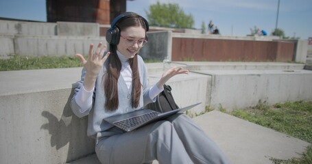 A hand-held shot of a young female student in casual clothes, glasses, and headphones making a video call on a laptop while sitting on a concrete bench in an urban park. © Anatoly Repin