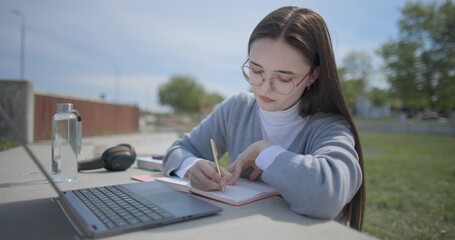 A hand-held shot of a young female student in casual clothes and glasses using a laptop and taking notes with a pen in a notebook in an urban park. © Anatoly Repin