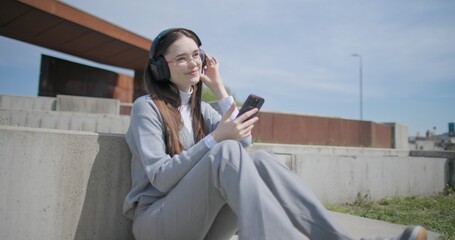 A hand-held shot of a young female student in casual clothes, glasses and headphones using a mobile phone while sitting on a concrete bench in an urban park. © Anatoly Repin