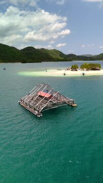Aerial view of a traditional wooden fishing platform floating on turquoise water near Gili Bedis island and lush green hills in Sekotong, West Nusa Tenggara, Indonesia.