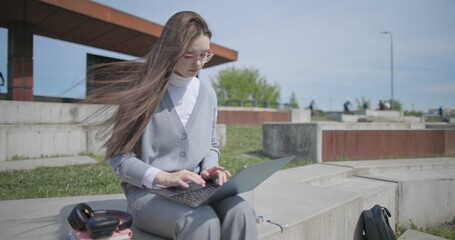 A hand-held shot of a young female student in casual clothes wearing glasses sitting on a concrete bench in an urban park on a sunny summer day, working on a laptop and typing on a keyboard. © Anatoly Repin