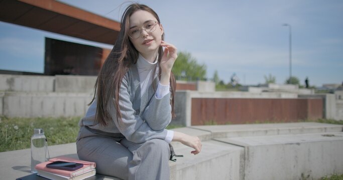 A handheld shot of a young female student in casual clothes sitting on a concrete bench in an urban park, enjoying a sunny summer day.