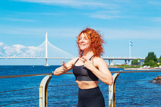 Energetic red-haired curly young woman in sportswear swinging her arms on the river embankment.