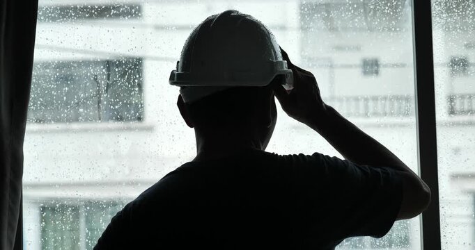 A silhouette of a male engineer stands inside a house, watching the rain fall through a window. The engineer is wearing a safety helmet.