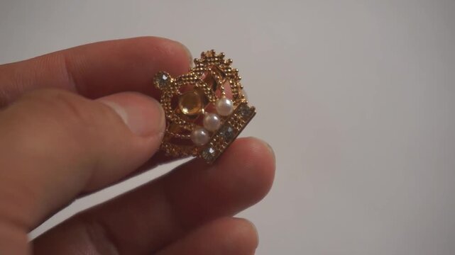 Close-up of human hand holding a golden crown-shaped brooch with pearls and gemstones, concept of luxury, royalty, elegance, and fashion accessories.