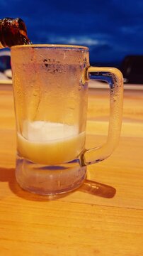 Vertical video. Cold Beer Pouring Into Frosted Glass Mug on Wooden Table Near Tropical Beach at Blue Hour, Refreshment and Vacation Lifestyle