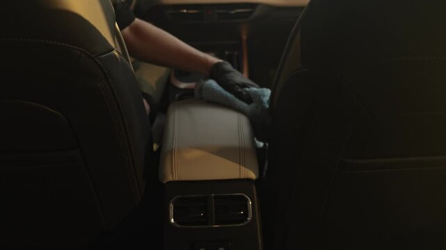 Gloved detailer wipes the armrest in the car, warm interior light, view from the back seat