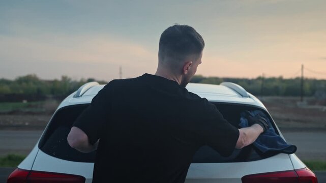 Man dries and polishes the rear trunk of his car outside in the evening, wiping away the remaining dirt with a towel.