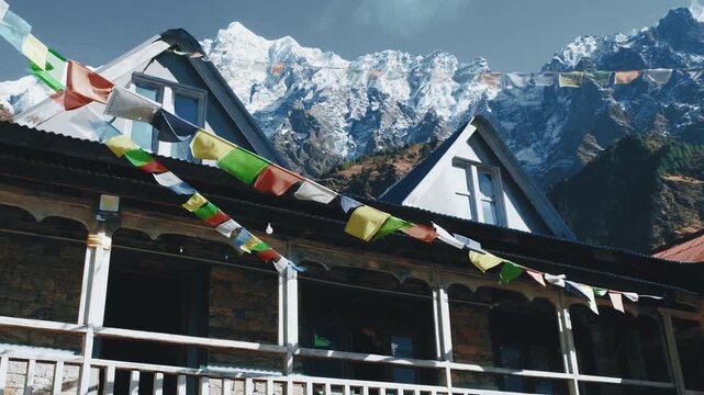  Colorful Tibetan prayer flags strung across a traditional Nepalese teahouse with snow-capped Manaslu mountains rising behind the rustic wooden lodge.