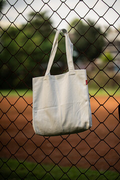 White tote bag hangs on chain-link fence. Soft lighting highlights fabric texture and simple design. Background shows grassy field and blurred trees. Casual