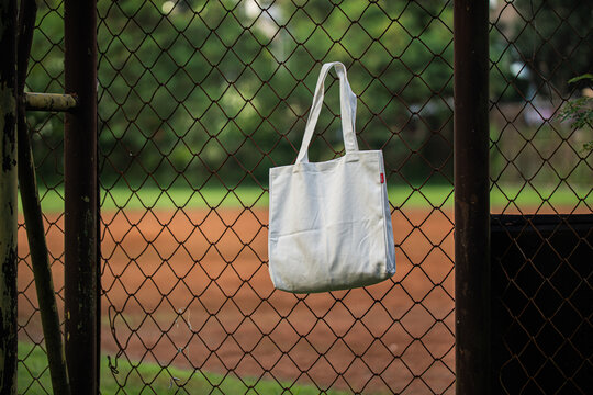White tote bag hangs on chain-link fence. Soft lighting highlights fabric texture and simple design. Background shows grassy field and blurred trees. Casual