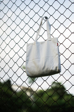 White tote bag hangs on chain-link fence. Soft lighting highlights fabric texture and simple design. Background shows grassy field and blurred trees. Casual