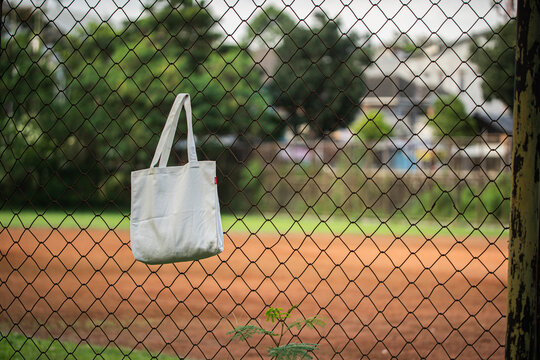 White tote bag hangs on chain-link fence. Soft lighting highlights fabric texture and simple design. Background shows grassy field and blurred trees. Casual