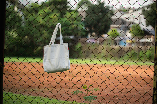 White tote bag hangs on chain-link fence. Soft sunlight highlights fabric's gentle folds. Background features blurred green grass and distant trees. No people visible, suggesting quiet, empty scene